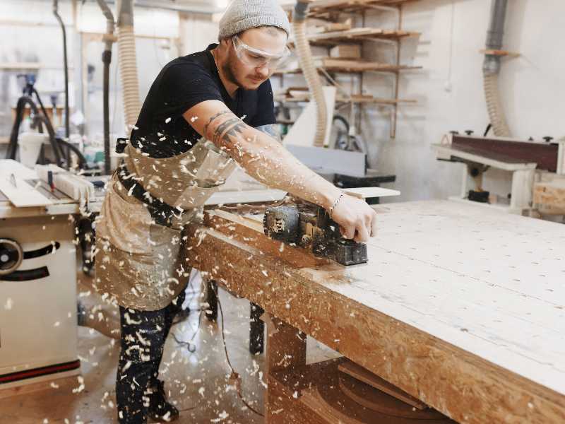 Woodworker using planer on plywood at workbench in woodshop, wood shavings flying